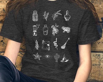 May include: Dark gray t-shirt featuring a white illustration of various mystical objects. The design includes a potion bottle, a snake, a star, a witch's hat, and other magical symbols. The shirt is made of a soft material.