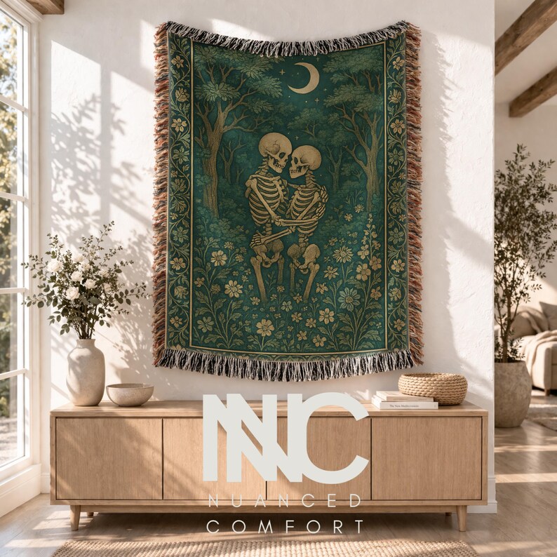 May include: A woven throw blanket featuring a dark teal forest scene with two skeletons embracing in a field of flowers under a crescent moon. The blanket has a decorative floral border and fringed edges on the top and bottom sides.
