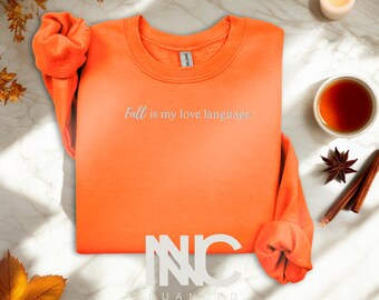Puede incluir: Sudadera naranja brillante con el texto "Fall is my love language". La imagen incluye una vela encendida, una taza de té, anís estrellado y palitos de canela, con temática otoñal.