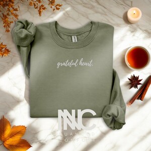 May include: An olive green sweatshirt with the words "grateful heart" in white script. The sweatshirt is displayed on a marble surface, with a lit candle, a cup of tea, and autumn decorations.