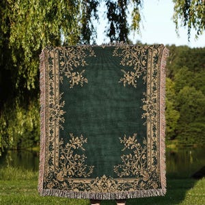May include: A dark green throw blanket with a beige floral pattern and fringe. The blanket is held up in front of a person standing on grass. The logo "INC Nuanced Comfort" is visible at the bottom.