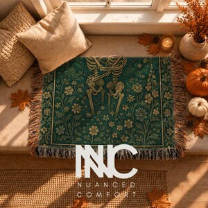 May include: A rectangular woven throw blanket featuring a dark teal background with a repeating pattern of cream-colored skeletons and floral botanical illustrations. The blanket has a fringed edge and displays the text NUANCED COMFORT in white.