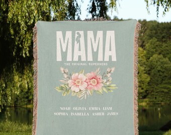 Può includere: Una coperta tessuta con uno sfondo blu chiaro e testo bianco che recita "Mama, The Original Superhero". La coperta presenta un design floreale con fiori rosa e foglie verdi. Il testo "Noah, Olivia, Emma, Liam, Sophia, Isabella, Asher, James" è stampato sotto il design floreale.