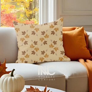 May include: A decorative pillow with a beige background and a repeating pattern of brown and gold maple leaves. An orange pillow is behind it. The pillow is on a white couch. The text "NUANCED COMFORT" is visible on the couch.