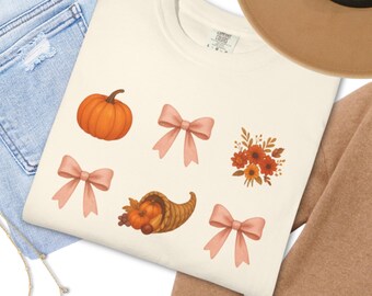 Puede incluir: Sudadera de color crema con gráficos temáticos de otoño. El diseño incluye una calabaza naranja, lazos rosas, una cornucopia y un arreglo floral en tonos naranja y rojo. La sudadera está hecha de un material suave.
