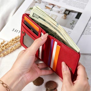 May include: A red bi-fold wallet filled with US $100 bills and various credit cards. The wallet is open, revealing multiple compartments and a snap closure. The image shows the wallet being held by a person.