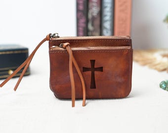 May include: A brown leather zipped pouch with a cross cut-out design. The pouch has a long strap that can be used as a wristlet or a shoulder strap.