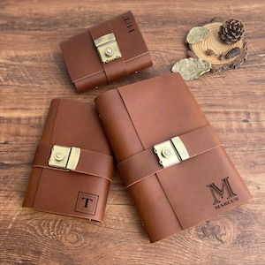 Könnte beinhalten: Drei braune Lederjournale mit Metallschließen und personalisierten Initialen. Die Journale haben ein rustikales Aussehen und Gefühl.