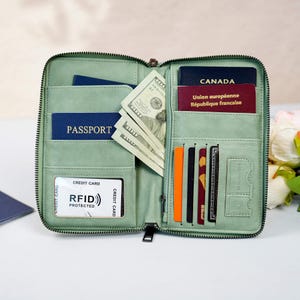 May include: A pale green leather travel wallet, open to show contents. Inside are a passport, Canadian passport, banknotes, and credit cards. The wallet has a zip closure and RFID protection.