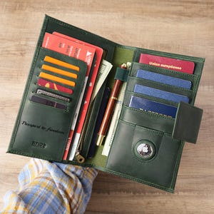 Könnte beinhalten: Offene grüne Reisebrieftasche aus Leder mit mehreren Kartenfächern, einer Reißverschlusstasche und Fächern für Dokumente. Die Brieftasche enthält einen Stift, Bargeld und einen kleinen AirTag. Der Text auf der Brieftasche lautet "Passport to freedom."