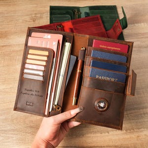 以下が含まれることがあります： 複数のカードスロット、ジッパー付きコンパートメント、ペンが付いた、開いた茶色の革製トラベルウォレット。ウォレットにはパスポートとカードが入っています。「Together We Explore the World」の文字が前面にエンボス加工されています。赤と緑の他のウォレットが背景に見えます。