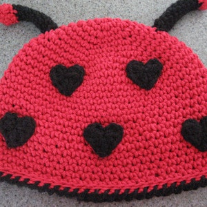 Puede incluir: Sombrero de mariquita de crochet rojo con corazones de fieltro negro y antenas de crochet negro.