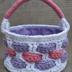 Peut inclure: Un panier en crochet avec une poignée rose et un motif rayé violet et blanc. Le panier est fait de fil et a une doublure en tissu.