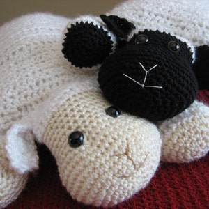 May include: Two crocheted sheep plushies, one white and one black, lying on a red surface. The white sheep has a beige face and black eyes. The black sheep has a white face and black eyes.