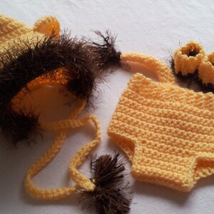 May include: A handmade crochet lion costume set in shades of yellow and brown. The set includes a hat with ears and a faux fur mane, a pair of booties, and a pair of shorts. The costume is designed for a baby.