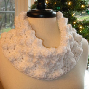May include: A white, crocheted cowl scarf with a textured, bubble-like pattern. The scarf is displayed on a mannequin, showcasing its circular design and cosy appearance. The background includes a Christmas tree.