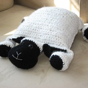 May include: A white crocheted sheep pillow with black crocheted facial features and legs. The sheep is lying on a light brown surface.