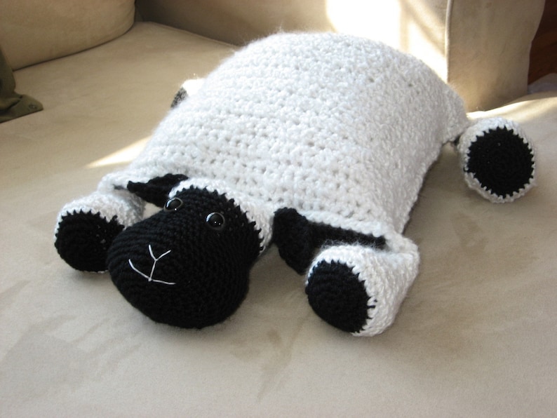 May include: A white crocheted sheep pillow with black crocheted facial features and legs. The sheep is lying on a light brown surface.