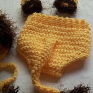 May include: A handmade, yellow crochet lion costume set for a baby, including a hat with a mane, diaper cover, and two ear covers. The mane and ear accents are brown.