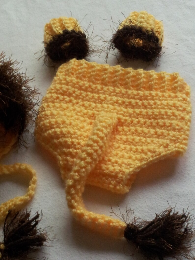 May include: A handmade, yellow crochet lion costume set for a baby, including a hat with a mane, diaper cover, and two ear covers. The mane and ear accents are brown.