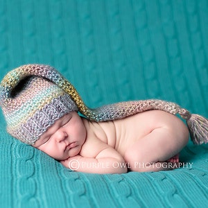 May include: A newborn baby wearing a multi-colored crocheted hat with a long tail, sleeping on a turquoise knitted blanket.