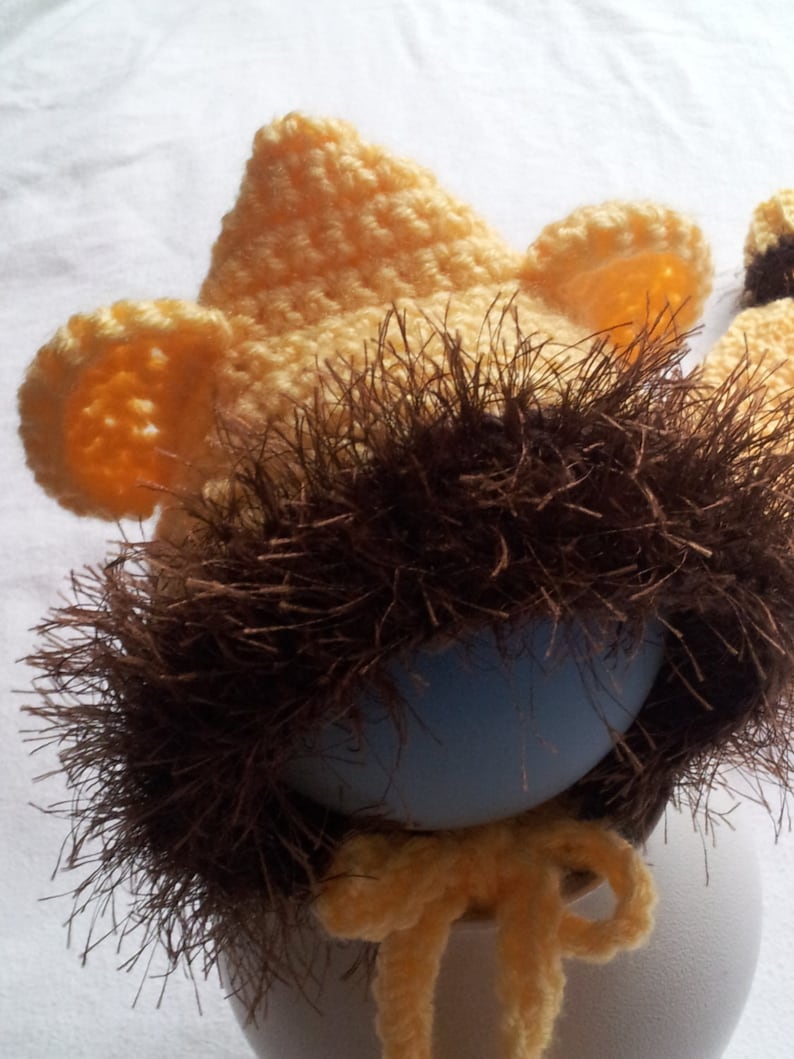 May include: A crocheted lion hat in shades of yellow and brown, designed to fit over an egg. The hat features a yellow crown, ear details, and a brown, shaggy mane. A yellow bow is tied at the base.