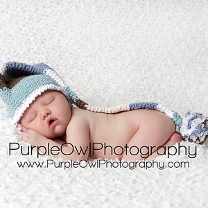 May include: A sleeping baby wearing a blue, brown, and white crocheted hat with a long tail and pom-pom. The baby is lying on a white, textured surface. The text "PurpleOwl Photography" and website address are visible.