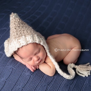 Puede incluir: Un bebé recién nacido con un gorro de crochet beige y blanco con una larga borla, durmiendo sobre una manta de punto azul. El bebé está acostado de lado con la cabeza apoyada en el brazo.