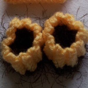 May include: Close-up of two handmade crochet sunflower appliques. The appliques have yellow petals and dark brown centres. They are surrounded by brown yarn, suggesting a rustic theme. The craft project is likely for embellishments.
