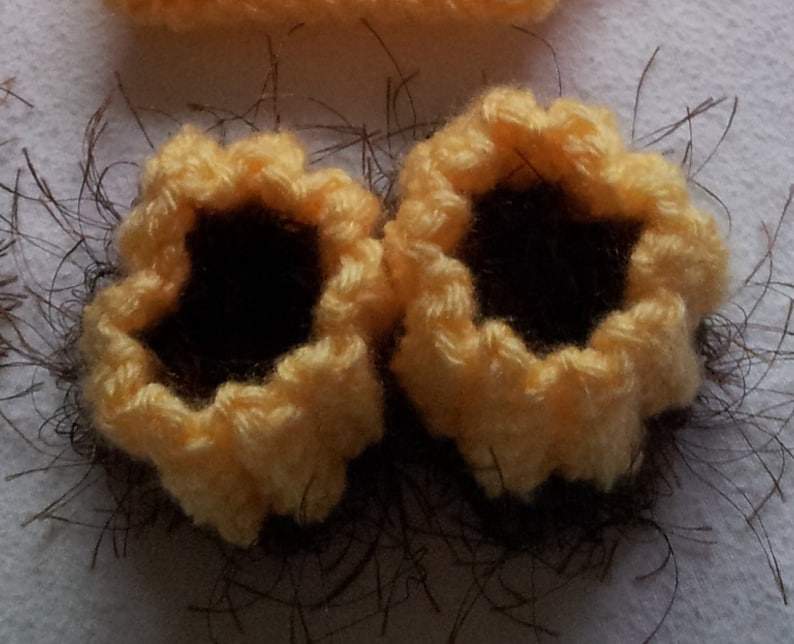 May include: Close-up of two handmade crochet sunflower appliques. The appliques have yellow petals and dark brown centres. They are surrounded by brown yarn, suggesting a rustic theme. The craft project is likely for embellishments.