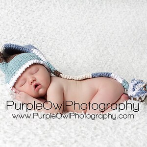 May include: A newborn baby wearing a blue, brown, and white crocheted hat with a long tail and pom-pom, sleeping peacefully on a white, textured surface. The image includes the text "PurpleOwlPhotography" and a website address.