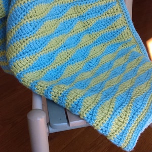 May include: A handmade crochet blanket in a blue and yellow wavy pattern. The blanket is draped over a light-colored wooden chair. The blanket's texture and color make it a cozy home decor item.