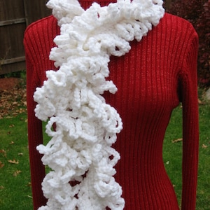 May include: A white crocheted scarf with a ruffled texture. The scarf is draped around a red turtleneck sweater.
