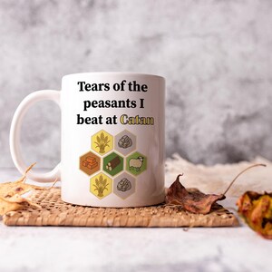 May include: White ceramic mug with the text "Tears of the peasants I beat at Catan" and game-themed graphics. The mug is on a woven coaster with dried leaves.