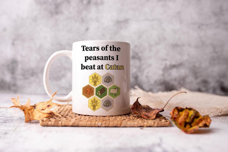 May include: White ceramic mug with the text "Tears of the peasants I beat at Catan" and game-themed graphics. The mug is on a woven coaster with dried leaves.