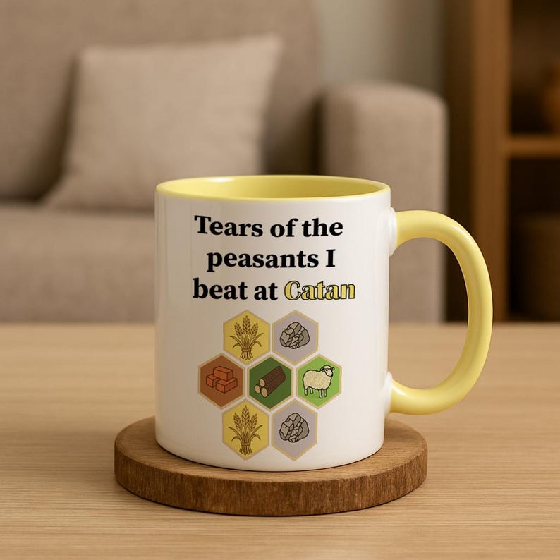 May include: White ceramic mug with a yellow interior and handle, featuring the text "Tears of the peasants I beat at Catan." The mug also displays game resource icons. The mug sits on a wooden coaster.