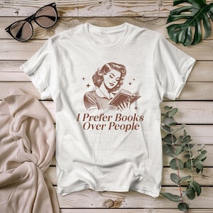 Peut inclure: T-shirt blanc avec une illustration brune d'une femme lisant un livre. Le texte "I Prefer Books Over People" est imprimé sous l'illustration.