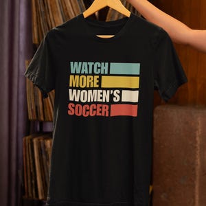Peut inclure: T-shirt noir avec le texte "WATCH MORE WOMEN'S SOCCER" en lettres majuscules colorées.