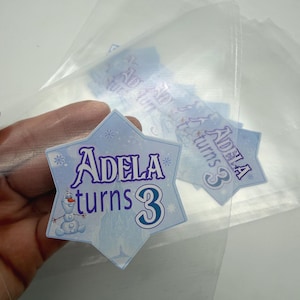 May include: A light blue star-shaped sticker with the text "Adela turns 3" in purple and blue. The sticker features a cartoon snowman and snowflake accents. Several transparent sheets are in the background.