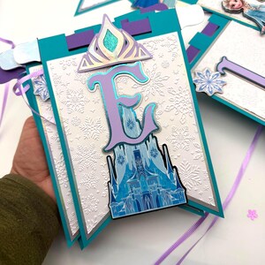May include: A banner with a teal border and a white background embossed with snowflakes. The banner features a purple letter "E" with a blue castle design, a silver crown, and a snowflake accent. Purple and white ribbons are attached.