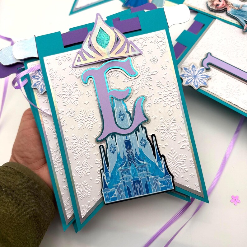 May include: A banner with a teal border and a white background embossed with snowflakes. The banner features a purple letter "E" with a blue castle design, a silver crown, and a snowflake accent. Purple and white ribbons are attached.