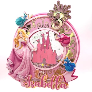 Puede incluir: Un adorno para pastel rosa y dorado con una princesa, un castillo y una cúpula llena de purpurina. El adorno también incluye un número 3 dorado y el nombre "Isabella".