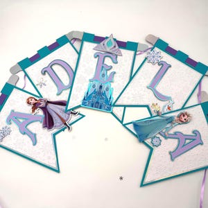 May include: A banner with the name "ADELA" in purple letters. Each letter is on a white pennant with a teal border. The banner features images of characters from the movie Frozen, including Elsa and Anna, and a castle.