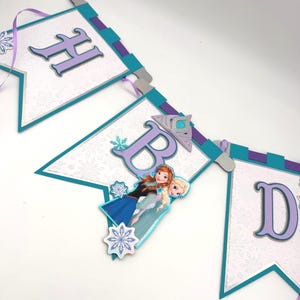 May include: A decorative banner with the letters "HBD" in purple, teal, and silver. The banner features images of the characters Anna and Elsa from the movie Frozen, with snowflake accents. The banner is strung with purple ribbon.