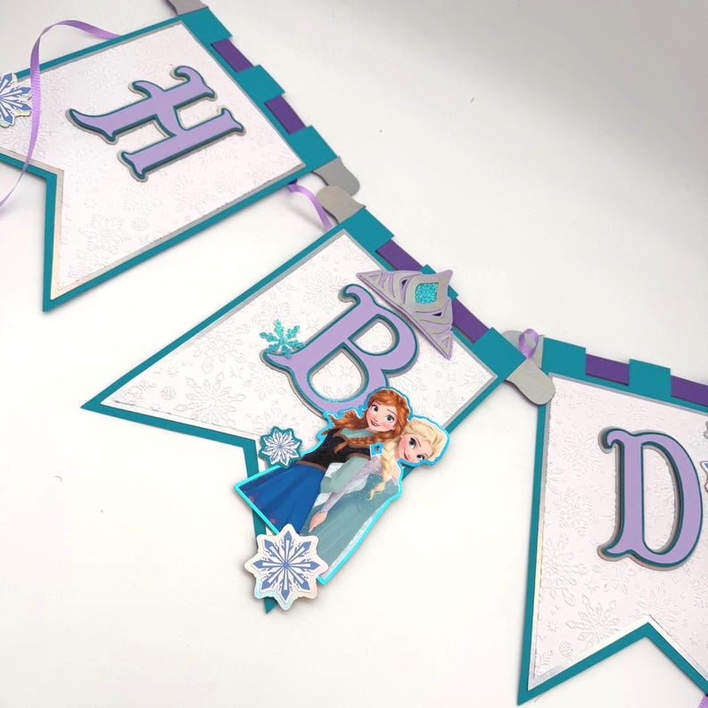 May include: A decorative banner with the letters "HBD" in purple, teal, and silver. The banner features images of the characters Anna and Elsa from the movie Frozen, with snowflake accents. The banner is strung with purple ribbon.