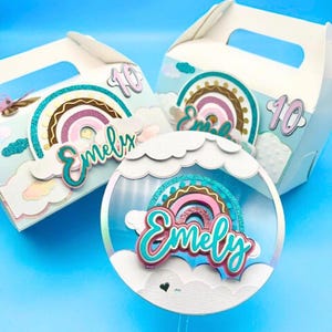 May include: Three white party favour boxes with rainbow and cloud decorations. Each box has a handle and a number 10. One box has a clear, round decoration with a rainbow and the name "Emely". The boxes are on a blue background.