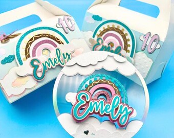 Könnte beinhalten: Drei weiße Partygeschenkboxen mit Regenbogen- und Wolkendekorationen. Jede Box hat einen Griff und die Zahl 10. Eine Box hat eine transparente, runde Dekoration mit einem Regenbogen und dem Namen "Emely". Auf blauem Hintergrund.