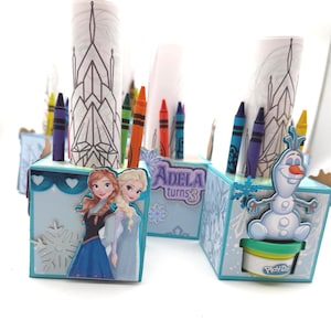 May include: Three Frozen-themed party favor boxes with crayons and coloring pages. Each box features Anna, Elsa, and Olaf characters. One box has the text "Adela turns 3". The boxes are light blue with snowflake and heart designs.