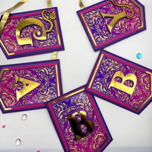 Op de afbeelding: Een verzameling decoratieve banners met gouden letters. Elke banner heeft een paarse en roze rand, met een gouden bloemenpatroon achtergrond. De gouden letters vallen op tegen de levendige achtergrond. Aan elke banner is een gouden lint bevestigd.