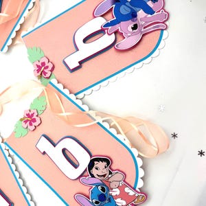 May include: A pink and blue birthday banner with the number '6', white borders, Stitch and Lilo characters, and hibiscus flower decorations. The banner has a scalloped edge and is strung with a sheer ribbon.